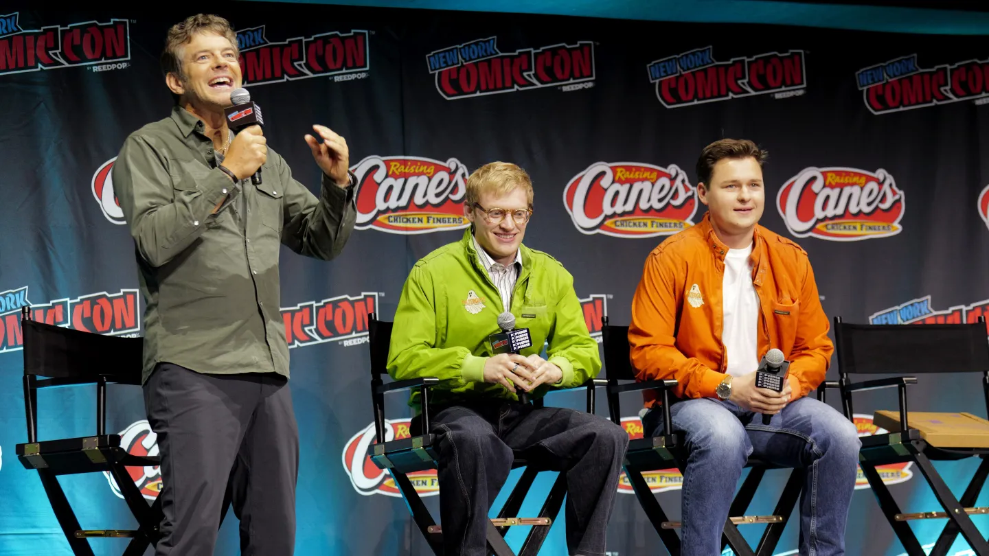 A man speaks and gestures beside two seated panelists at New York Comic Con, with event and sponsor logos behind them.